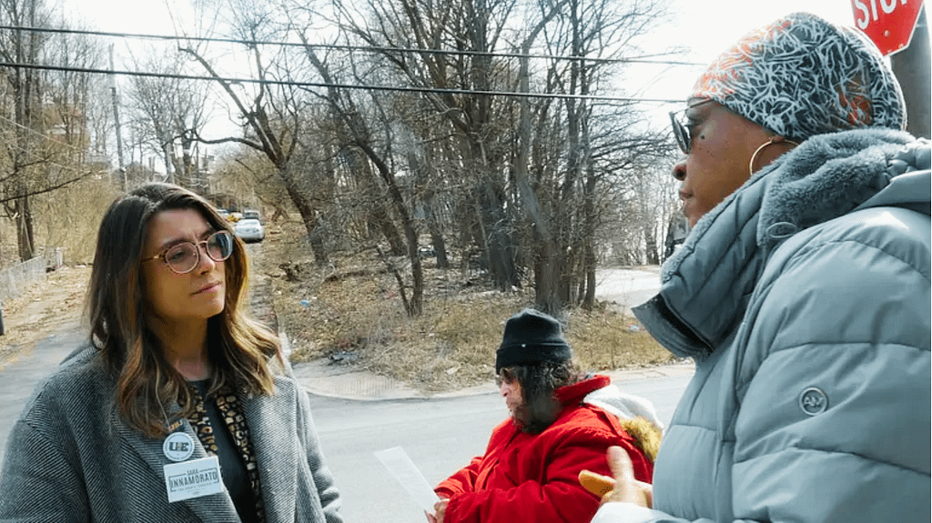 image description: Sara Innamorato (left) listens intently to Ms. Sherai Richardson (right), who is a participant in the fare pilot, at the bus stop in Lincolm-Lemington on a cold afternoon. Ms. Verna Johnson (center) sits near the stop waiting for the bus as well.