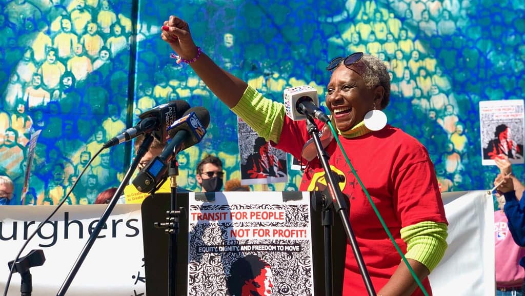 image description: PPT member Debra Green leads a rally for affordable fares. She is smiling and has her fist raised in the air. PPT Members stand behind her holding signs.