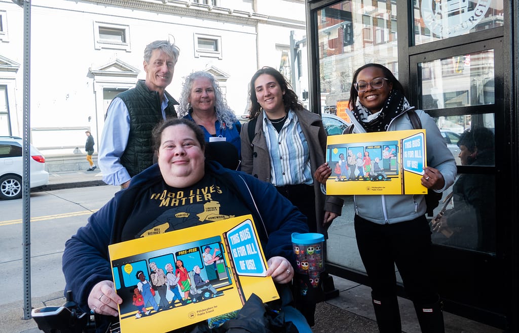 David Fawcett and intern Ally, standing at a bus shelter in Downtown, Pittsburgh and smiling with PPT Ride-along delegation. Alisa Grishman and Cheryl Stephens are holding signs from PPT saying “This Bus Is For All of Us!”