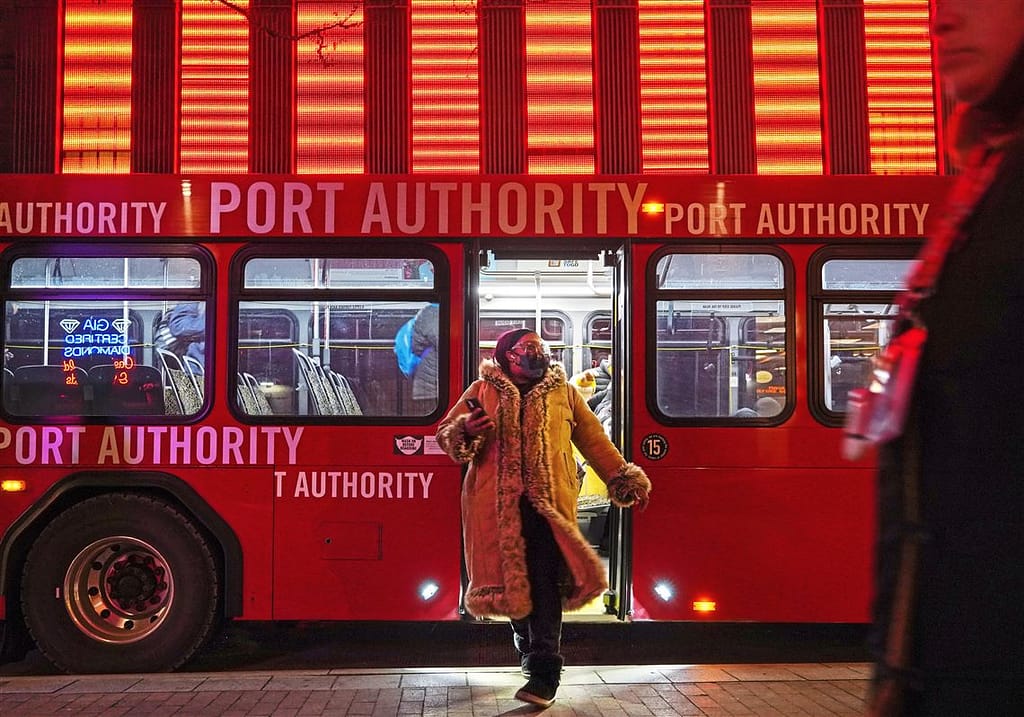 Photo from the Pittsburgh Post-Gazette of a woman walking off of a red Port Authority bus downtown at Wood Street Station. It is night time and the neon above the station glows red as the breeze blows the front of her long coat.