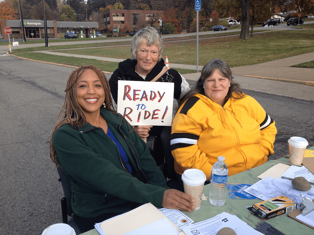 image of three PPT members canvassing at a table. One is holding a handwritten sign that reads “Ready to Ride!”
