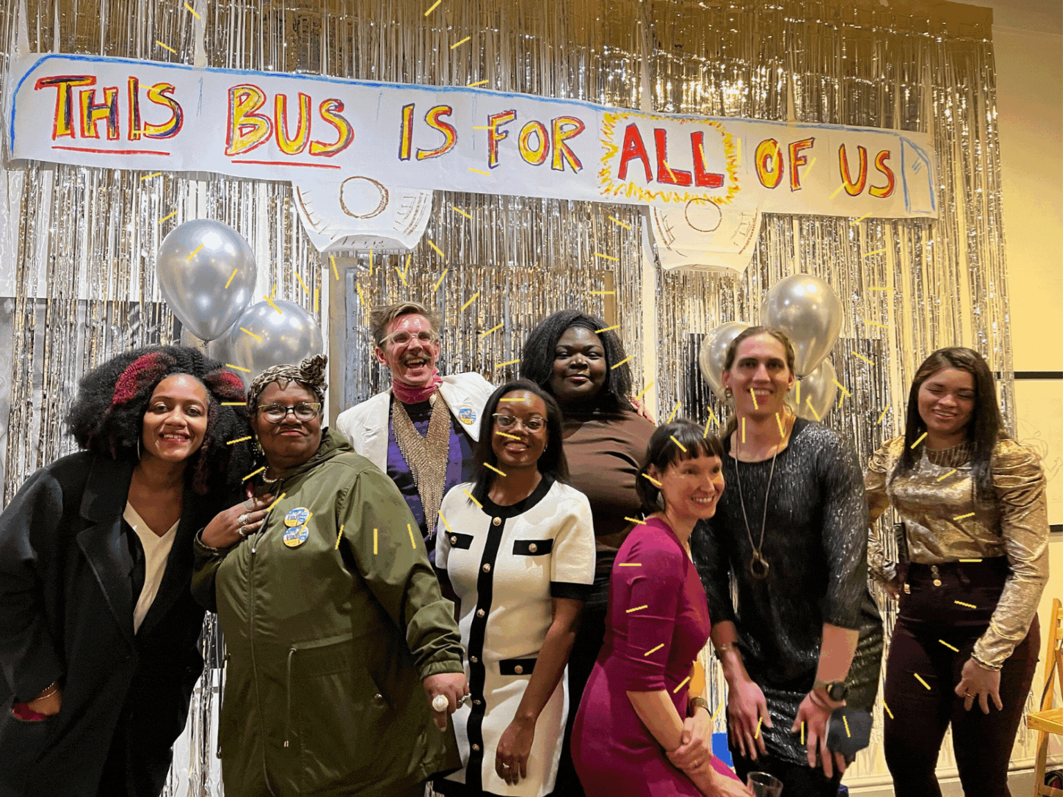 Image Description: PPT Members post at the photo booth. Everyone is smiling and wearing wonderful outfits, bright colors and fun, shiny clothing. The photo booth backdrop is silver streamers, silver ballons and a handmade sign that reads: “THIS BUS IS FOR ALL OF US”.