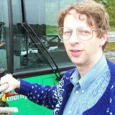 Image Description: Stu Strickland, a white man with brown hair, glasses, and a blue sweater, is looking at the camera while standing and holding a bicycle handle in front of a bus.
