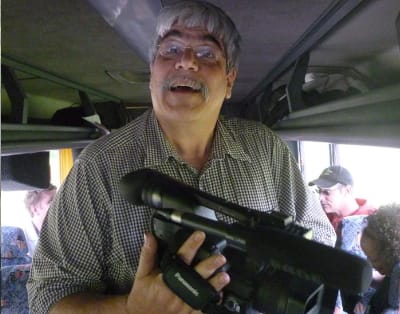 Image description: Dean Mougianis, a white man with white hair and glasses wearing a plaid shirt, holds a video camera on a bus during a past rally day in Harrisburg. Other rally attendees are seated in the background.