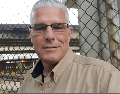 Photo of Kevin, white man, with short white hair and glasses. He’s wearing his Port Authority uniform and is standing under a bridge. A chainlink fence is immediately behind him, and transit tracks are in the background.
