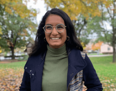 Image description: I am an Indian-American woman with shoulder length hair. In this photo, I am wearing a green turtleneck shirt with a navy jacket, glasses, and gold hoop earrings. I am standing in front of fall scenery with colorful trees behind me.