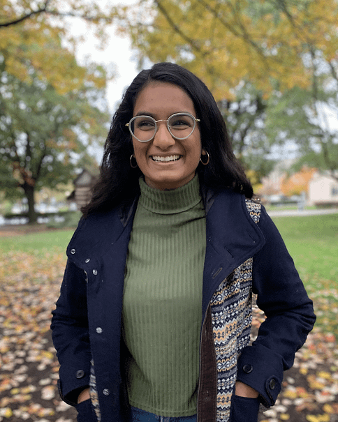 Image description: I am an Indian-American woman with shoulder length hair. In this photo, I am wearing a green turtleneck shirt with a navy jacket, glasses, and gold hoop earrings. I am standing in front of fall scenery with colorful trees behind me.