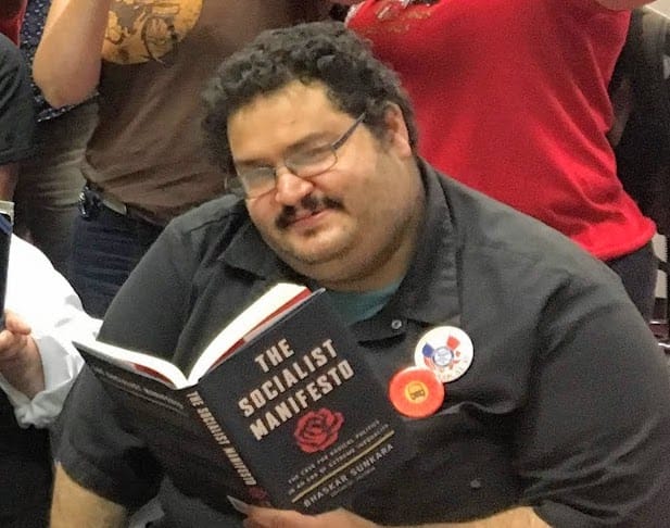 Image description: Andrew Hussein, a middle-aged White male with a mustache and black hair reading a book on socialism and other liberal topics