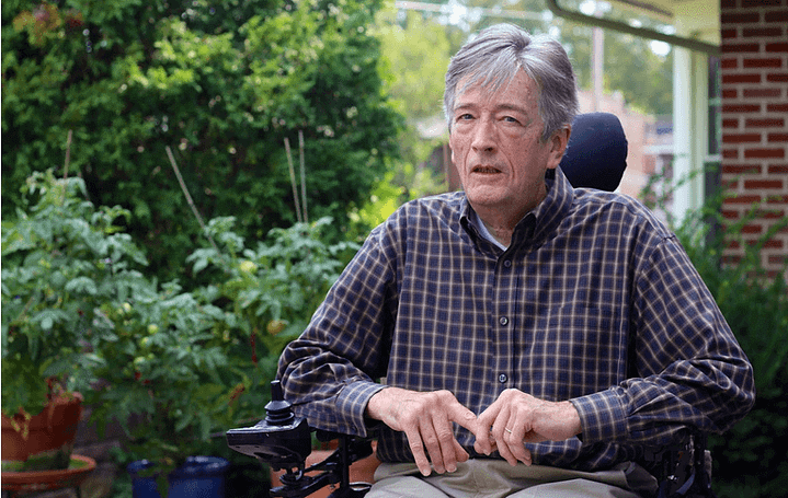 Image Description: Paul W. O’Hanlon, white man with white hair and a plaid shirt, sits in his power wheelchair in a garden. Tomato plants and the corner of a red brick house are in the background.