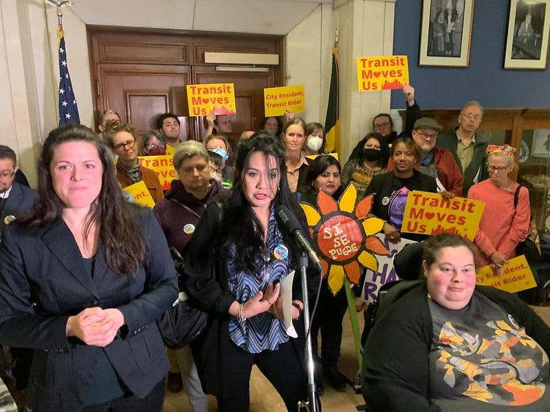 Image Description: PPT Member Lorena speaks at a PPT rally and release of our “Representing Our Routes” report in the Spring of 2023. She is flanked by an ASL interpreter and dozens of PPT members holding red and yellow signs.